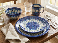 A place setting on a rustic wooden table featuring a matching ceramic dinnerware set with blue and green floral mandala patterns. The set includes a large plate, a smaller plate, a bowl, a mug, silver cutlery, and a beige linen napkin.