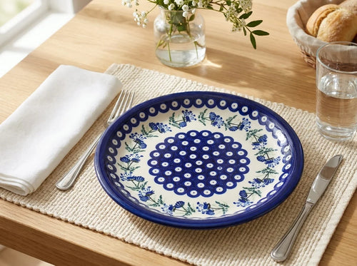 Polish pottery plate in Sweet Annie pattern on woven placemat with silverware, a vase of flowers, and a basket of bread on a wood table.