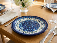 A round ceramic table plate with a detailed blue and white mandala pattern, set on a wooden dining table with silverware, a folded napkin, and a glass vase of white flowers.