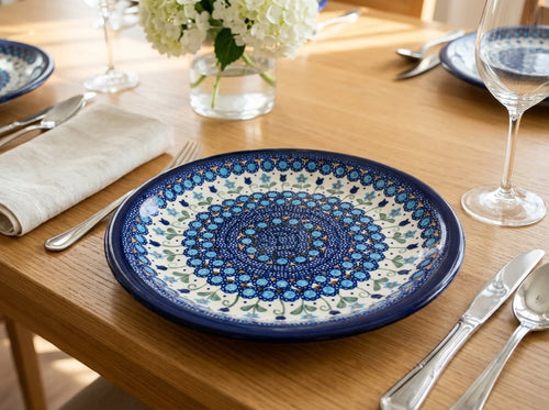 A round ceramic table plate with a detailed blue and white mandala pattern, set on a wooden dining table with silverware, a folded napkin, and a glass vase of white flowers.