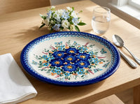 A round ceramic plate with a traditional Polish floral pattern in blue, yellow, and green, set on a light wood dining table next to a folded white napkin, silver spoon, glass of water, and a small flower bouquet.