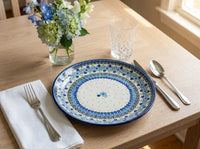 Polish pottery plate in Eliza pattern on wood table with silverware, a crystal glass, and a vase of flowers.