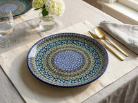 A round ceramic plate with an intricate blue, green, and gold mandala pattern, styled on a beige linen table runner with gold cutlery and a glass vase of white flowers.