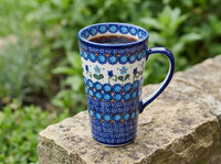 A tall ceramic mug with a blue and white floral pattern sits on a stone surface outdoors with green foliage blurred in the background.