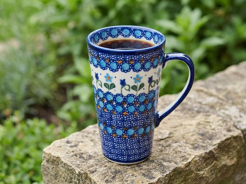 A tall ceramic mug with a blue and white floral pattern sits on a stone surface outdoors with green foliage blurred in the background.