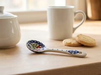 A white ceramic tea spoon with blue floral patterns and a small red flower detail, resting on a light wooden table next to a white mug and cookies.