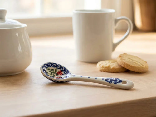 A white ceramic tea spoon with blue floral patterns and a small red flower detail, resting on a light wooden table next to a white mug and cookies.