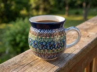 A ceramic coffee mug with a colorful floral pattern and dark blue rim, filled with coffee and resting on a wooden deck railing with a blurred green outdoor background.
