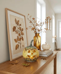 A styled autumn interior scene showing a wooden console table with a framed botanical print, a gold vase with dried branches, a carved pumpkin lantern with a tea light, a small white pumpkin, and books.