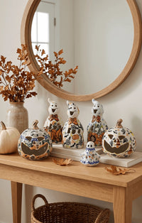 A styled entryway console table featuring a set of ceramic Halloween decorations, including three tall ghost figurines, two carved jack-o'-lantern pumpkins, and a small ghost, arranged under a large round wooden mirror.