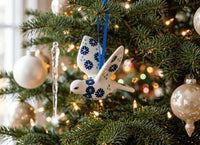 A white ceramic bird ornament with blue floral patterns hangs on a decorated Christmas tree with green branches, warm string lights, and white and gold baubles.