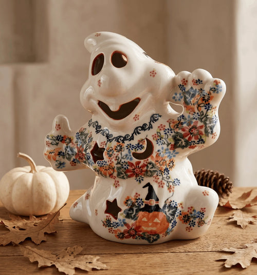 A ceramic ghost figurine decorated with autumn floral patterns, featuring a smiling face, waving hands, and a pumpkin-and-moon design on a rustic wooden surface with a small pumpkin and leaves.
