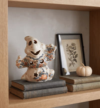 A ceramic ghost figurine decorated with orange, blue, and black floral patterns sits on a stack of vintage books on a wooden shelf, next to a small pumpkin and a framed botanical print.