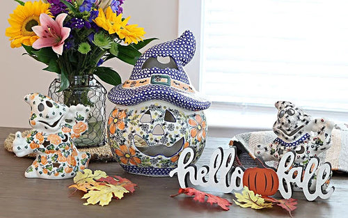 A set of ceramic Halloween decorations displayed on a wooden table in front of a window. The set includes a central witch hat jack-o'-lantern, a small ghost figurine, a Dalmatian dog figurine, a 'hello fall' sign, and a small pumpkin accent, all decorated with orange, yellow, and green floral patterns.