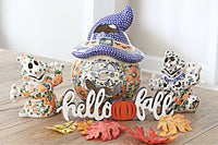 Three hand-painted ceramic fall decorations arranged on a wooden tabletop: a witch hat gingerbread man, a ghost with pumpkins, and a Dalmatian-style skeleton, with a 'hello fall' sign and autumn leaves in front.