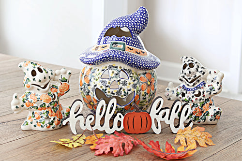 Three hand-painted ceramic fall decorations arranged on a wooden tabletop: a witch hat gingerbread man, a ghost with pumpkins, and a Dalmatian-style skeleton, with a 'hello fall' sign and autumn leaves in front.