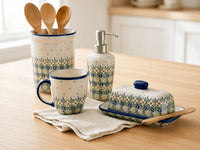 A coordinated kitchenware set on a light wood table, including a floral-patterned mug, soap dispenser, butter dish with lid, and a tall spoon holder with wooden spoons. The pieces have a cream base with blue and green floral motifs and trim.