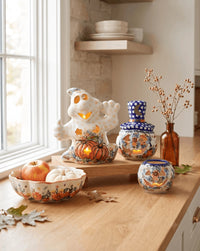 A styled autumn kitchen counter display featuring a white ceramic ghost candle holder with a glowing pumpkin base, a blue-and-white patterned ceramic jar, a matching small container, and a brown glass vase with dried stems, alongside a bowl of pumpkins and fall leaves.