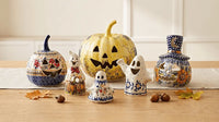 A set of hand-painted ceramic Halloween decorations, including a jack-o'-lantern, three ghosts, and a small top hat, arranged on a wooden table with autumn leaves and acorns.