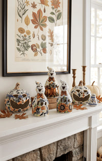 A set of ceramic Halloween ghost pumpkins with autumn floral patterns displayed on a white fireplace mantel below a framed botanical print.