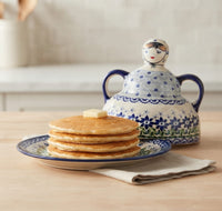 A stack of golden pancakes with a pat of butter on top sits on a blue-and-white patterned plate, with a matching blue-and-white lidded pitcher behind it on a light wood surface.