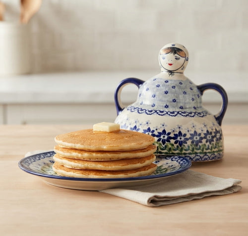 A stack of golden pancakes with a pat of butter on top sits on a blue-and-white patterned plate, with a matching blue-and-white lidded pitcher behind it on a light wood surface.