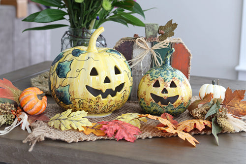Two decorative hand-painted pumpkins with carved jack-o'-lantern faces sit on a burlap runner with autumn leaves and pinecones on a wooden table. A small orange pumpkin, a white acorn, and a rustic sign are arranged around them, with a vase of green leaves in the background.