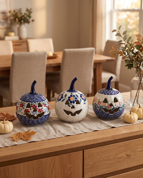 Three decorative ceramic pumpkins with hand-painted floral and jack-o'-lantern designs are displayed on a wooden table runner in a warm, dining-room setting.