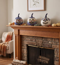 Three decorative ceramic pumpkins with blue and floral patterns are displayed on a wooden fireplace mantel in a cozy living room setting.