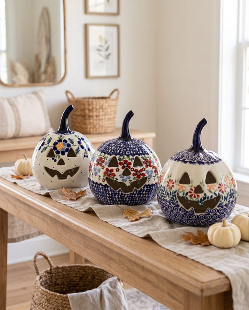 Three hand-painted ceramic jack-o'-lantern pumpkins with carved faces are displayed on a wooden table runner in a cozy, sunlit room. The pumpkins feature intricate floral and geometric patterns in blue, white, orange, and brown, surrounded by small decorative pumpkins and autumn leaves.