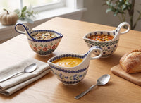 Three ceramic gravy boats with floral patterns are arranged on a wooden table, each filled with a different soup. A folded beige napkin with a spoon, a loaf of bread on a cutting board, and a window with plants are visible in the background.