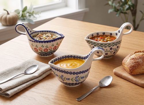 Three ceramic gravy boats with floral patterns are arranged on a wooden table, each filled with a different soup. A folded beige napkin with a spoon, a loaf of bread on a cutting board, and a window with plants are visible in the background.