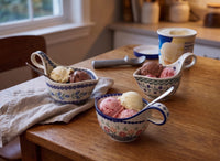 Three floral ceramic ice cream bowls filled with scoops of vanilla and chocolate ice cream on a wooden table, with an ice cream container and scoop in a softly lit kitchen setting.