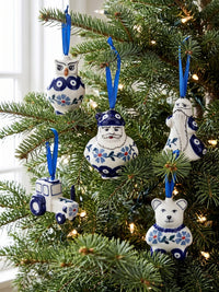 Five hand-painted ceramic Christmas tree ornaments in blue and white, including an owl, Santa, a tractor, a bearded man, and a polar bear, hanging on a green Christmas tree with warm string lights.