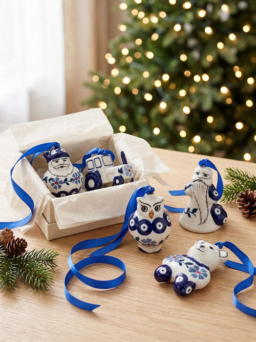 A set of ceramic Christmas ornaments in a white and blue glaze, displayed next to their gift box on a wooden table with a blurred Christmas tree in the background. The ornaments include a Santa Claus, a snowman, a snowman with a beard, an owl, and a polar bear, all with blue ribbons.