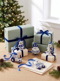 A set of ceramic Christmas ornament shapes styled on a wooden table with wrapped gift boxes, pine branches, and a blurred Christmas tree in the background. The ornaments include an owl, a tractor, a snowman, a bear, and a bird, decorated in a classic blue and white pattern.