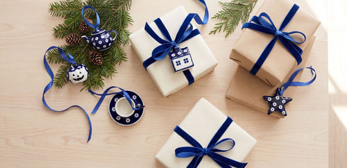 Three wrapped gift boxes on a light wood surface, decorated with navy blue ribbons and holiday ornaments including pine branches, a teapot ornament, a small cup ornament, and a star ornament.