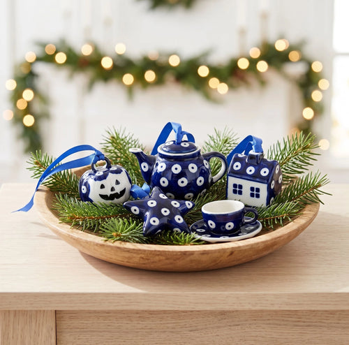 A set of ceramic Christmas tea-time accessories, including a teapot, cup and saucer, house-shaped teapot, and jack-o'-lantern ornament, all in a deep blue and white pattern, arranged in a wooden bowl with green pine branches on a light wood table.