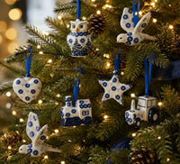 A close-up of a decorated Christmas tree with green branches, warm white string lights, and blue-and-white ceramic ornaments shaped like an owl, birds, a heart, a star, a train, and a tractor.