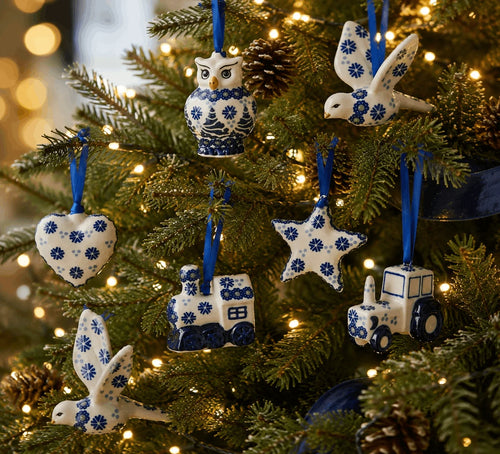 A close-up of a decorated Christmas tree with green branches, warm white string lights, and blue-and-white ceramic ornaments shaped like an owl, birds, a heart, a star, a train, and a tractor.