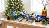 A set of blue and white ceramic Christmas ornaments shaped like a star, owl, snowman, and small vehicles, displayed on a wooden table with pine branches, a blurred Christmas tree, and an amber glass bottle in the background.