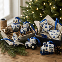 Blue and white ceramic Christmas ornament set displayed on a wooden table, including a bird, owl, tractor, train, and star shapes, with some ornaments in a gift box and others arranged beside pinecones and evergreen branches.