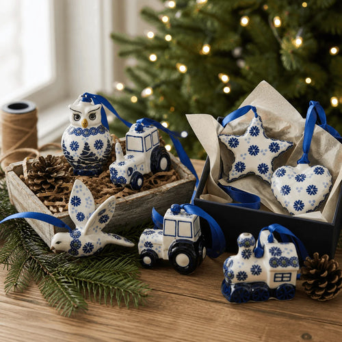 Blue and white ceramic Christmas ornament set displayed on a wooden table, including a bird, owl, tractor, train, and star shapes, with some ornaments in a gift box and others arranged beside pinecones and evergreen branches.