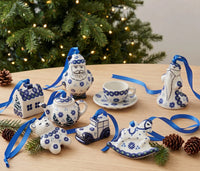 A collection of ceramic Christmas ornaments shaped like a house, Santa, teapot, teacup, rocking horse, snowman, and a train, decorated in a classic blue and white Polynesian style with blue ribbons, arranged on a wooden table with pinecones and a blurred Christmas tree in the background.