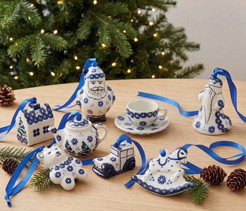 A collection of ceramic Christmas ornaments shaped like a house, Santa, teapot, teacup, rocking horse, snowman, and a train, decorated in a classic blue and white Polynesian style with blue ribbons, arranged on a wooden table with pinecones and a blurred Christmas tree in the background.