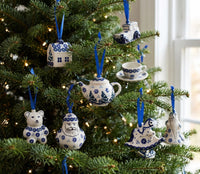 Blue and white ceramic Christmas tree ornaments shaped like a teapot, teacup, bear, Santa Claus, and other holiday figures hanging on a decorated Christmas tree with warm string lights.