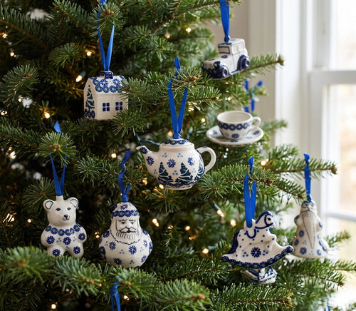 Blue and white ceramic Christmas tree ornaments shaped like a teapot, teacup, bear, Santa Claus, and other holiday figures hanging on a decorated Christmas tree with warm string lights.