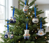 A decorated Christmas tree with six hand-painted ceramic ornaments in blue, white, and orange floral patterns, including an owl, bear, tractor, ghost, and jack-o'-lantern shapes, hanging in the green branches of the tree.