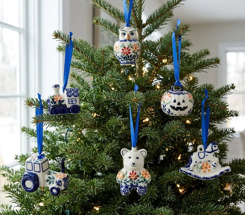 A decorated Christmas tree with six hand-painted ceramic ornaments in blue, white, and orange floral patterns, including an owl, bear, tractor, ghost, and jack-o'-lantern shapes, hanging in the green branches of the tree.