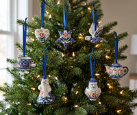 A set of hand-painted ceramic Christmas ornaments hanging on a decorated Christmas tree. The ornaments include a teacup and saucer, a bird, a Santa Claus figure, a teapot, a flower, and a bird with a floral pattern, all decorated in a classic blue, white, and red Polish pottery style with blue ribbons.